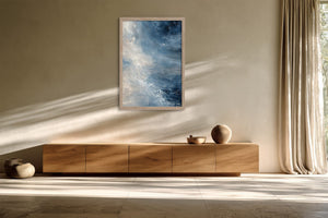Framed blue abstract wall art above a low wooden sideboard beside long curtains.