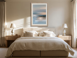 Framed blue-grey abstract seascape hangs above a bed between matching bedside lamps.