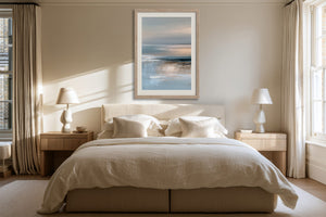 Framed blue-grey abstract seascape hangs above a bed between matching bedside lamps.