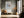 Framed abstract seascape above a bench with an orange cushion in a sunlit hallway.