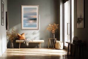 Framed abstract seascape above a bench with an orange cushion in a sunlit hallway.