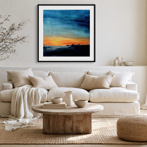 Black-framed abstract sunset print above a white sofa with pillows and a round wood coffee table.