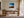 Black-framed abstract sunset print above a wooden desk beside tall windows.