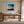 Black-framed abstract sunset print above a wooden desk beside tall windows.