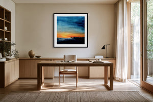 Black-framed abstract sunset print above a wooden desk beside tall windows.