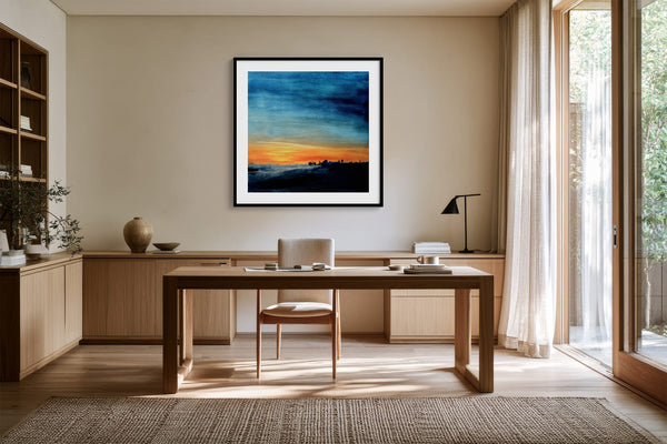 Black-framed abstract sunset print above a wooden desk beside tall windows.
