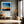 Black-framed abstract sunset print above a bench with an orange cushion and dried branches.