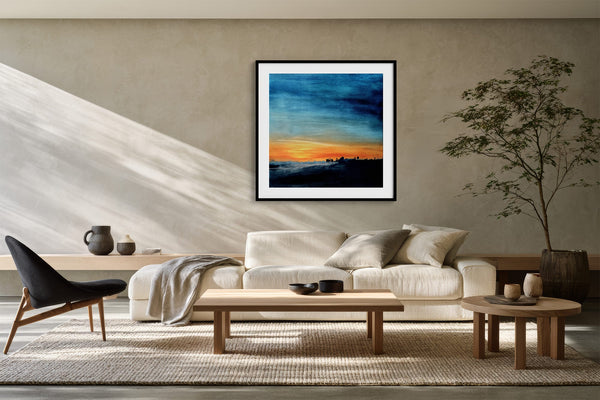 Black-framed abstract sunset print above a cream sofa, wood tables, and a potted tree.