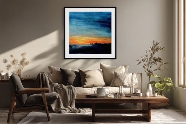Black-framed abstract sunset print above a cream sofa, wood coffee table, and leafy plants.
