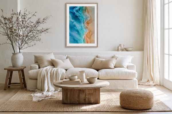 Light wood-framed ocean-and-sand artwork above a cream sofa, rustic table, and woven ottoman.