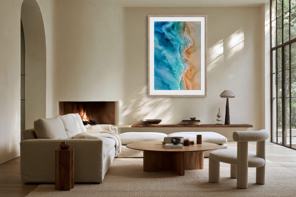 White-framed coastal artwork above a low wood console in a sunlit seating area with fireplace.