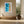 Framed turquoise-and-sand aerial artwork above a built-in bench beside large windows and ceramic vessel.