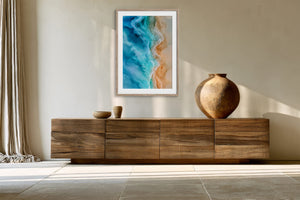 Framed aerial ocean artwork displayed above a long wooden sideboard with a round ceramic vase.