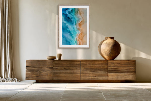 Framed aerial ocean artwork displayed above a long wooden sideboard with a round ceramic vase.
