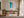 Framed coastal aerial artwork above a wooden desk in a bright workspace with open glass doors.