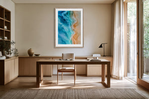 Framed coastal aerial artwork above a wooden desk in a bright workspace with open glass doors.