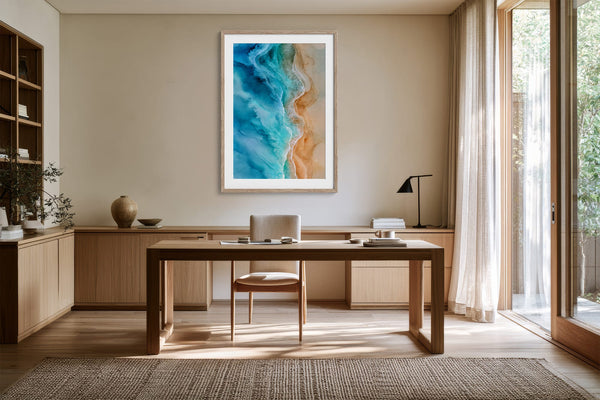 Framed coastal aerial artwork above a wooden desk in a bright workspace with open glass doors.
