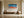 Framed turquoise-wave and orange-shore artwork above a wooden bench beside a large ceramic vessel.