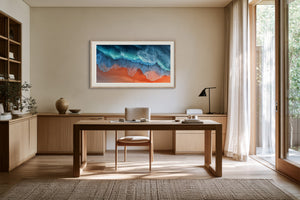 Framed aerial wave artwork displayed above a wooden desk in a sunlit workspace.
