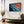Ember Tide artwork in a thin wood frame above a bench and gold vase of branches.