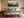 Large framed watercolor of layered pine trees above a beige bed with a bench.