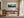 Framed watercolor of misty pine trees above a wooden desk near tall glass doors.