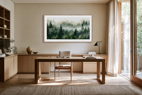 Framed watercolor of misty pine trees above a wooden desk near tall glass doors.