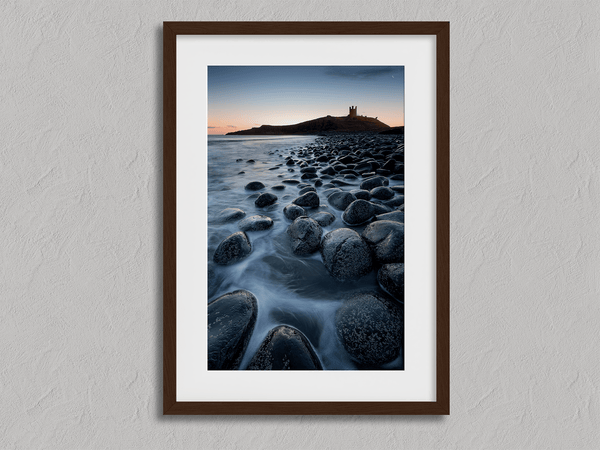 Framed fine art print of Northumbrian castle ruins at sunrise, with glowing rocks, tranquil sea, and lingering moonlight in soft dawn hues.