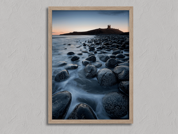 Framed fine art print of Northumbrian castle ruins at sunrise, with glowing rocks, tranquil sea, and lingering moonlight in soft dawn hues.