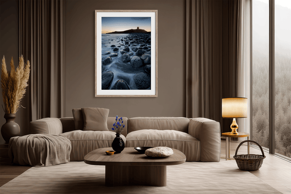 Framed fine art print of Northumbrian castle ruins at sunrise, with glowing rocks, tranquil sea, and lingering moonlight in soft dawn hues.