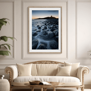Framed fine art print of Northumbrian castle ruins at sunrise, with glowing rocks, tranquil sea, and lingering moonlight in soft dawn hues.
