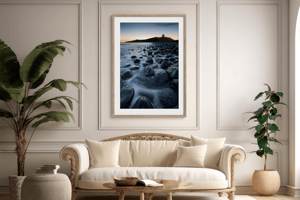 Framed fine art print of Northumbrian castle ruins at sunrise, with glowing rocks, tranquil sea, and lingering moonlight in soft dawn hues.