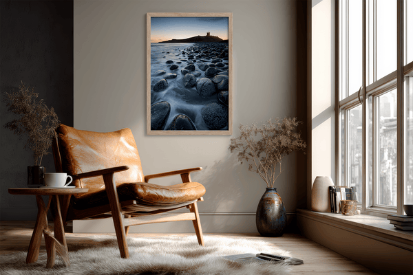 Framed fine art print of Northumbrian castle ruins at sunrise, with glowing rocks, tranquil sea, and lingering moonlight in soft dawn hues.