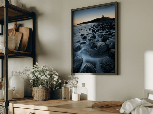 Framed fine art print of Northumbrian castle ruins at sunrise, with glowing rocks, tranquil sea, and lingering moonlight in soft dawn hues.