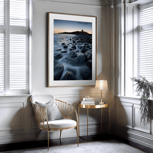 Framed fine art print of Northumbrian castle ruins at sunrise, with glowing rocks, tranquil sea, and lingering moonlight in soft dawn hues.