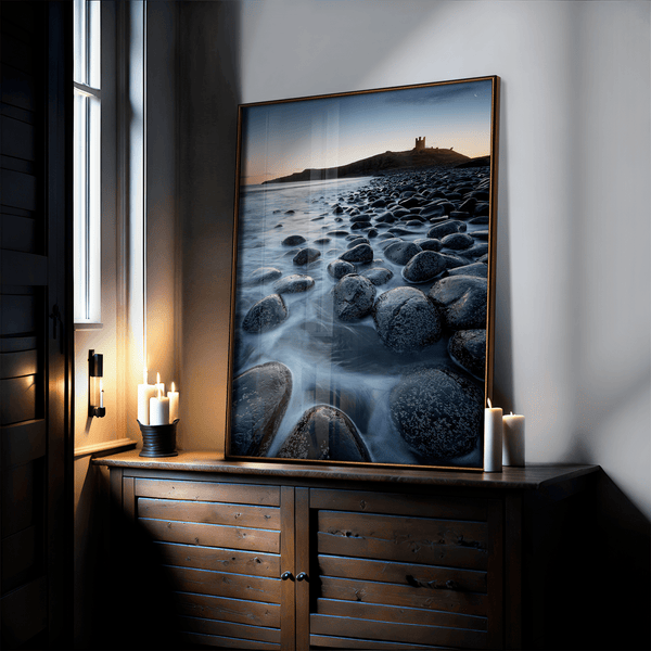 Framed fine art print of Northumbrian castle ruins at sunrise, with glowing rocks, tranquil sea, and lingering moonlight in soft dawn hues.
