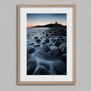 Framed fine art print of Northumbrian castle ruins at sunrise, with glowing rocks, tranquil sea, and lingering moonlight in soft dawn hues.
