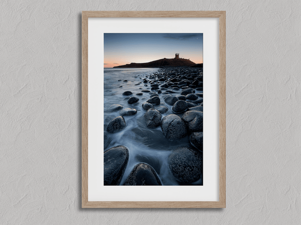 Framed fine art print of Northumbrian castle ruins at sunrise, with glowing rocks, tranquil sea, and lingering moonlight in soft dawn hues.