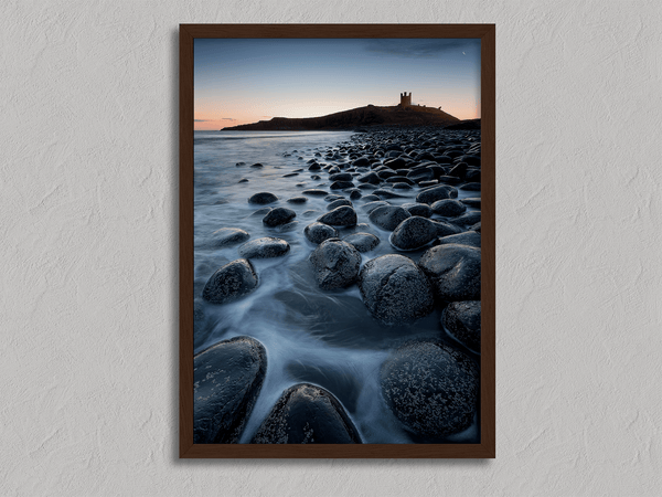 Framed fine art print of Northumbrian castle ruins at sunrise, with glowing rocks, tranquil sea, and lingering moonlight in soft dawn hues.