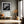 Black-framed lighthouse print above a bench with an orange pillow in a sunlit hallway.