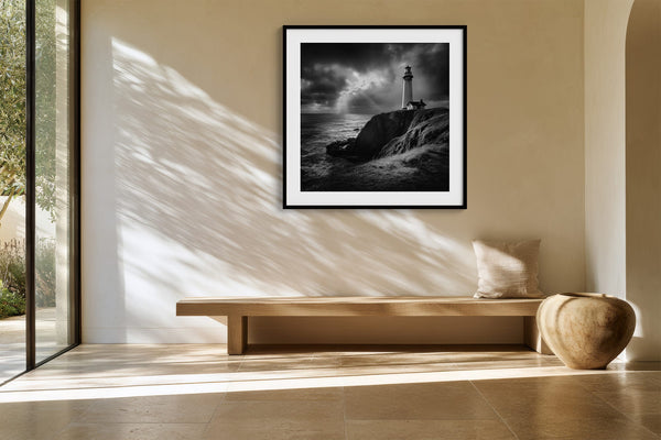 Black-framed lighthouse print above a long wood bench in a sunlit space with glass doors.