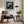 Black-framed lighthouse print above a brown armchair beside a floor lamp and bright windows.