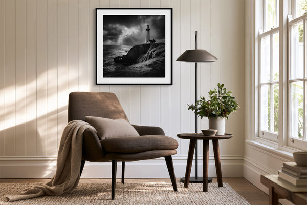 Black-framed lighthouse print above a brown armchair beside a floor lamp and bright windows.