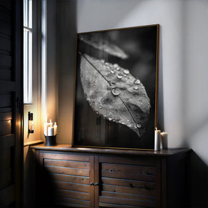 Black-and-white dew-covered leaf wall art in a thin dark frame, leaning on a wooden cabinet beside candles.