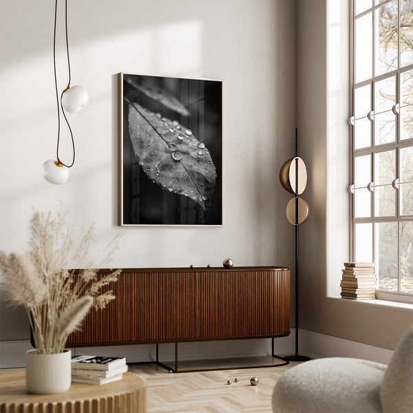 Black-and-white leaf wall art in a light frame above a wooden sideboard beside a large window.