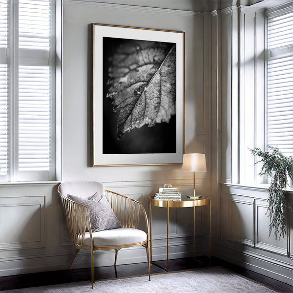 Black-and-white leaf artwork in a thin gold frame with white mat above a chair.