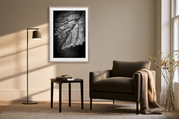 White-matted leaf print in a light wood frame above an armchair and side table.