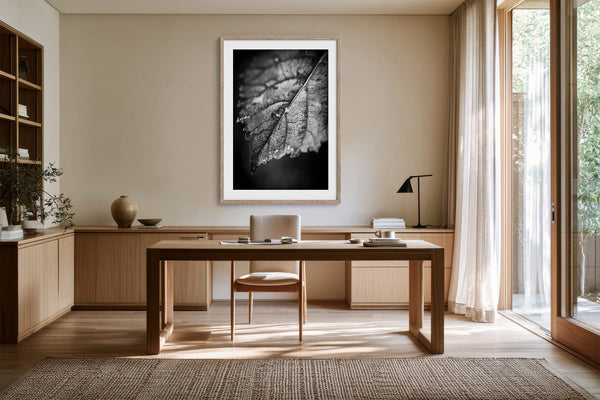 White-matted black-and-white leaf print in a light wood frame above a wooden desk.