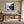 Black-framed Mistwater Valley print above a wooden desk near tall glass doors.