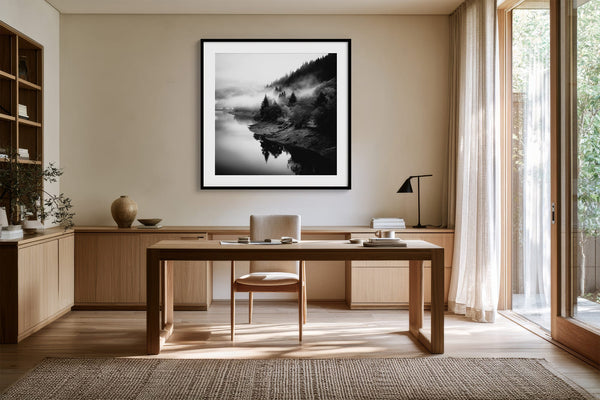 Black-framed Mistwater Valley print above a wooden desk near tall glass doors.
